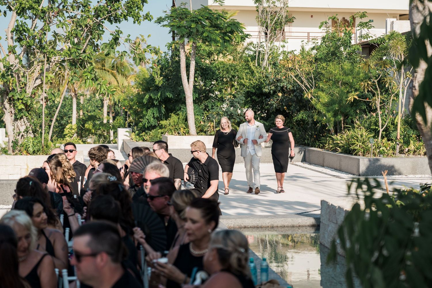 Guests dressed in black gather outdoors at an elegant venue surrounded by lush tropical greenery for a formal event.