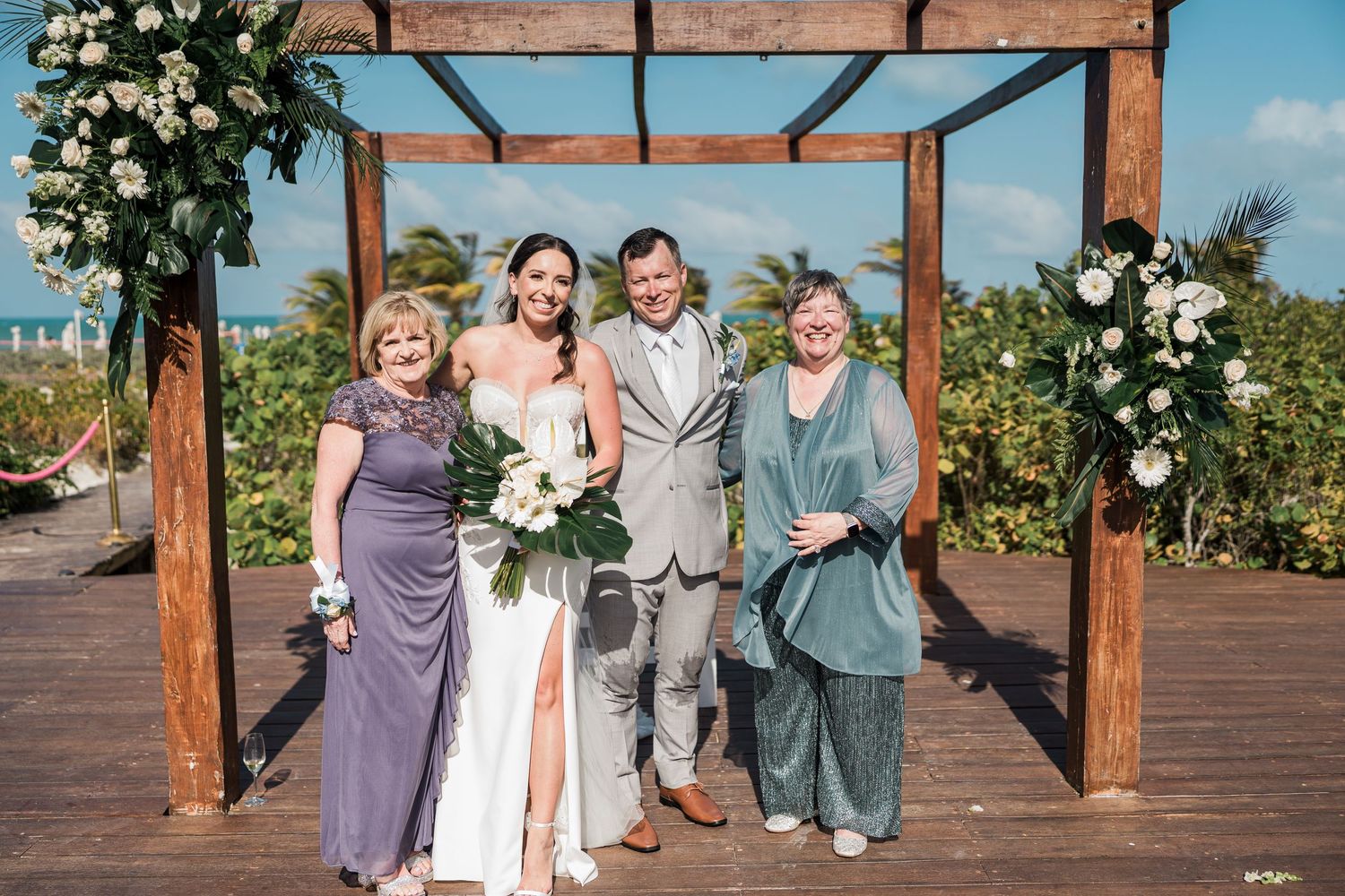Bride and groom pose with two guests under a wooden arch adorned with white flowers at a beachside wedding ceremony.