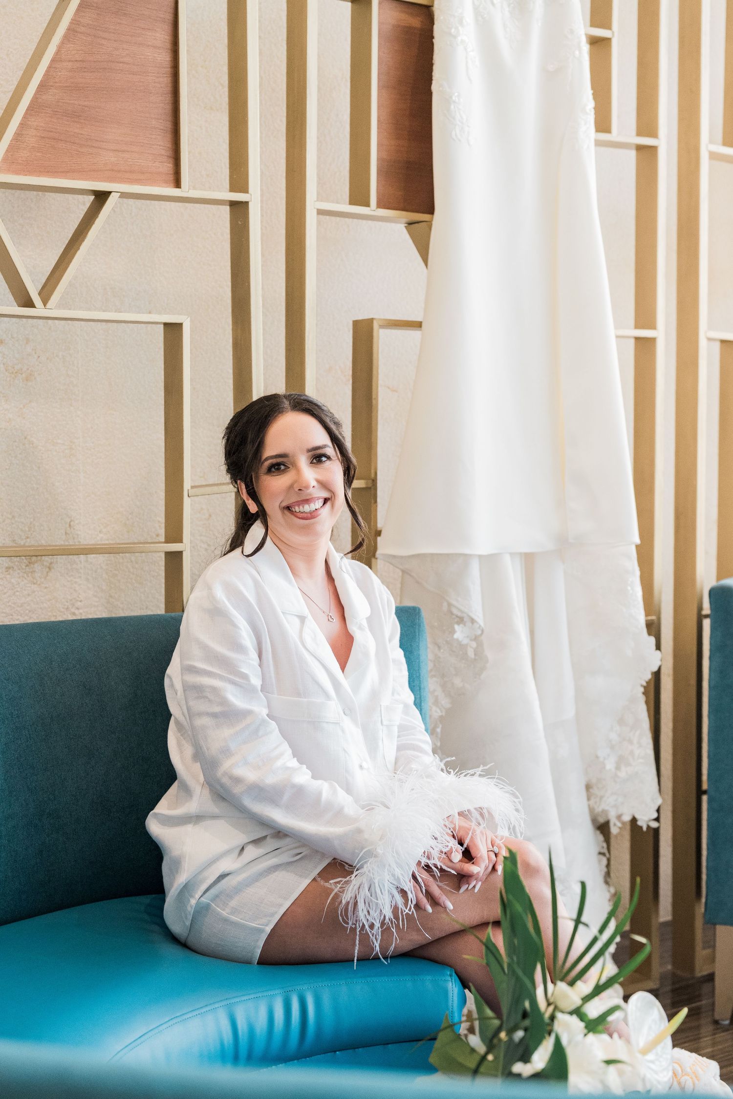 Smiling bride in white feather-trimmed robe sitting on blue velvet chair beside hanging wedding dress.
