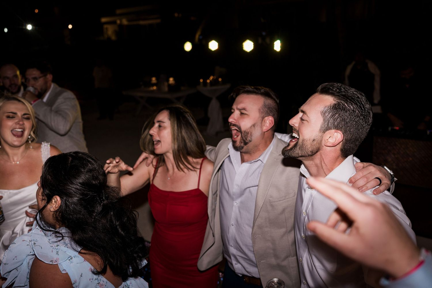 Group of friends laughing and dancing together at an outdoor evening wedding reception celebration.