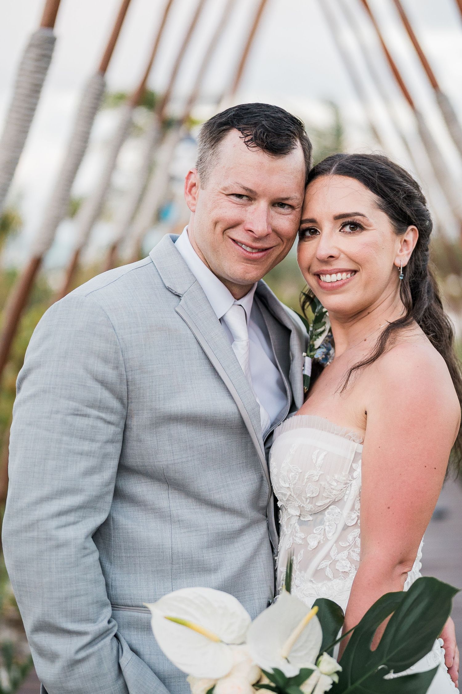 Smiling bride and groom pose together outdoors, bride holding white tropical bouquet, groom in light gray suit.