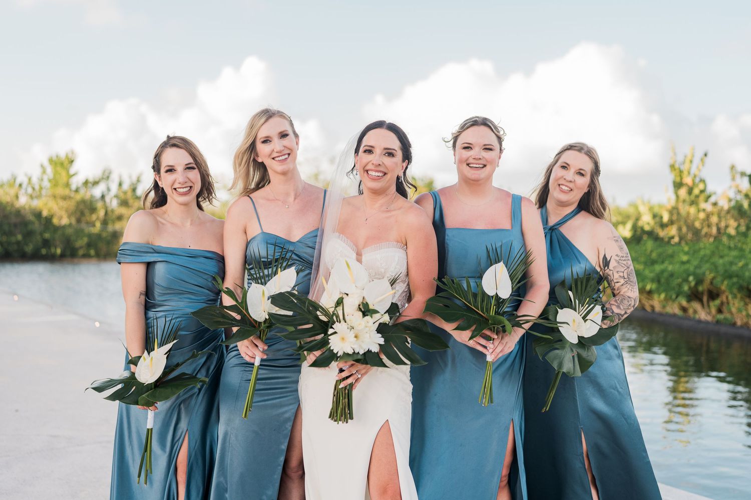 Bride in white gown with bridesmaids in blue satin dresses holding tropical bouquets outdoors near water.