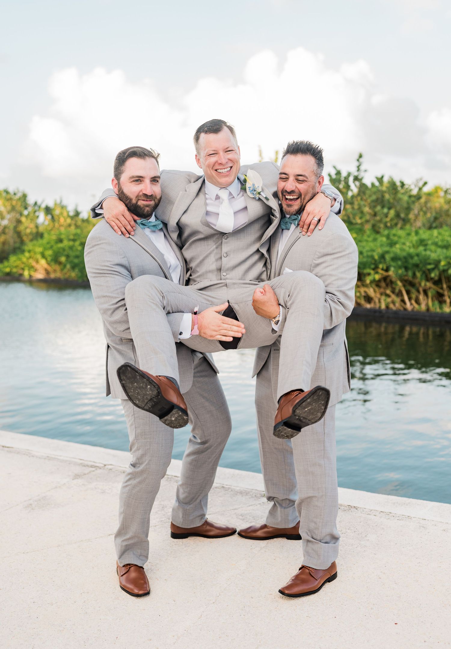 Three groomsmen in gray suits laughing as two carry the groom outdoors near a waterfront at a wedding.