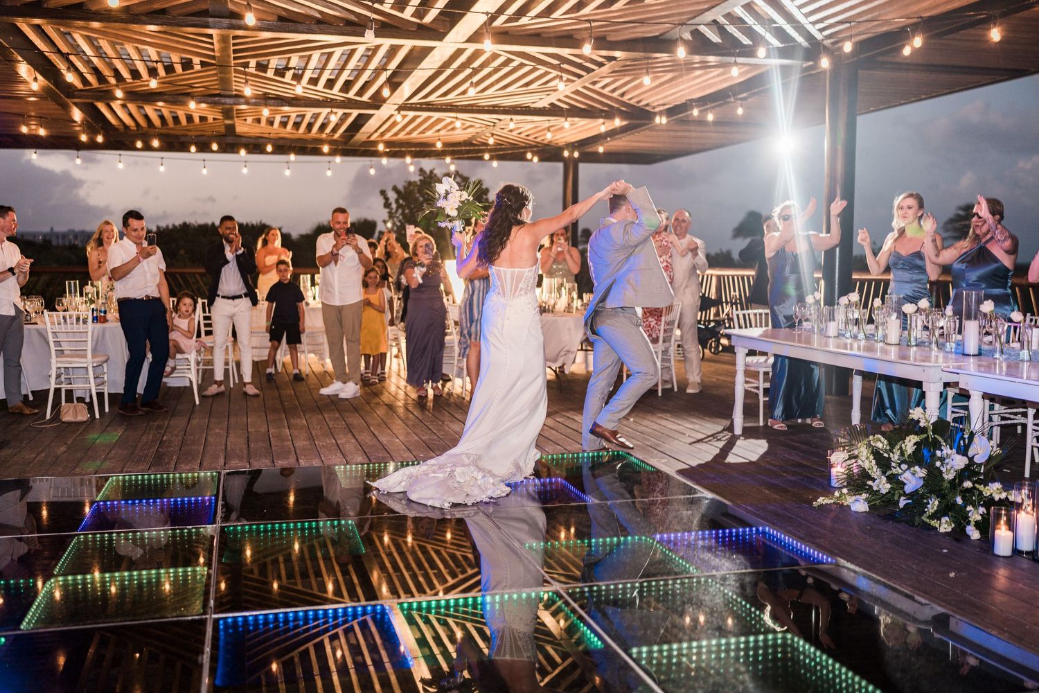 grand palladium costa mujeres Bride and groom share first dance on mirrored floor at elegant outdoor wedding reception with string lights.