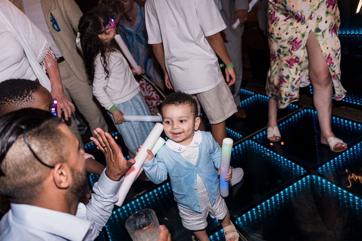 Toddler in light blue outfit dancing on LED dance floor at a party, surrounded by adults and children.