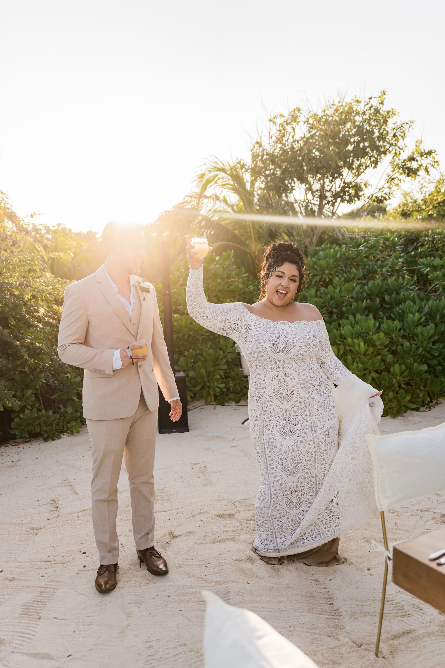 Xcaret Arte Wedding - Bride in lace gown and groom in tan suit celebrate on sandy beach at sunset during outdoor wedding ceremony.