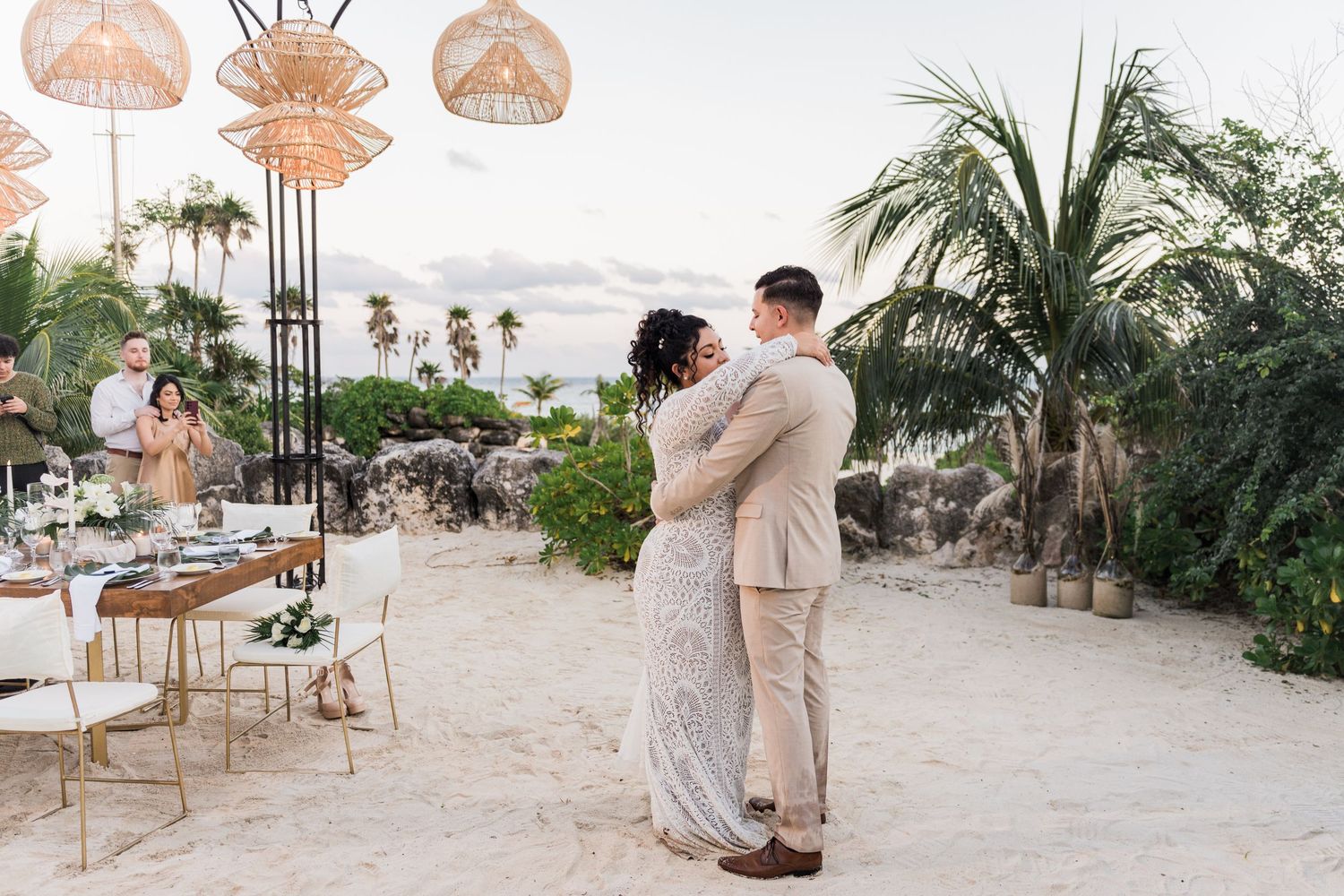 Xcaret Arte Wedding - Couple shares first dance on sandy beach wedding venue with wicker pendant lights and tropical palms at sunset.