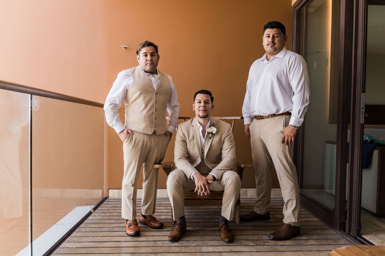 Xcaret Arte Wedding - Groom seated with two groomsmen in tan suits and white shirts on a balcony at a wedding.