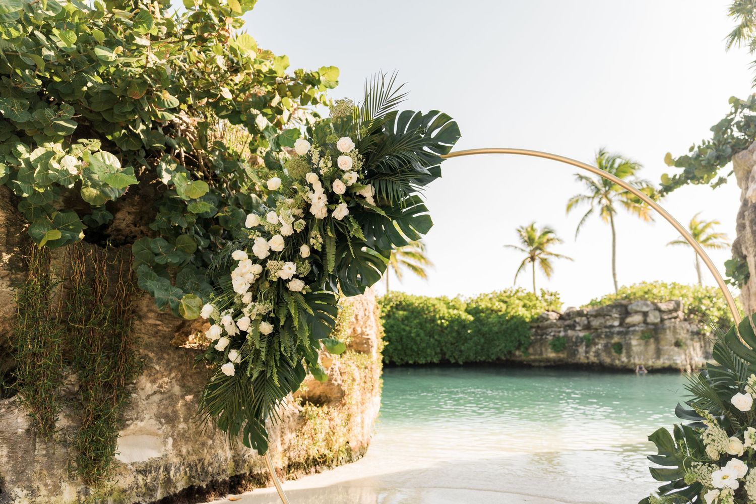 Xcaret Arte Wedding - Tropical wedding arch adorned with white flowers and monstera leaves overlooking a turquoise lagoon with palm trees.
