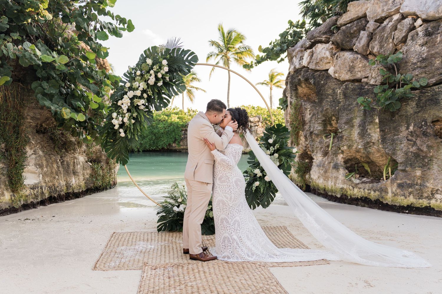 Xcaret Arte Wedding - Bride and groom share a romantic kiss under a floral arch at a tropical beach wedding venue with palm trees.