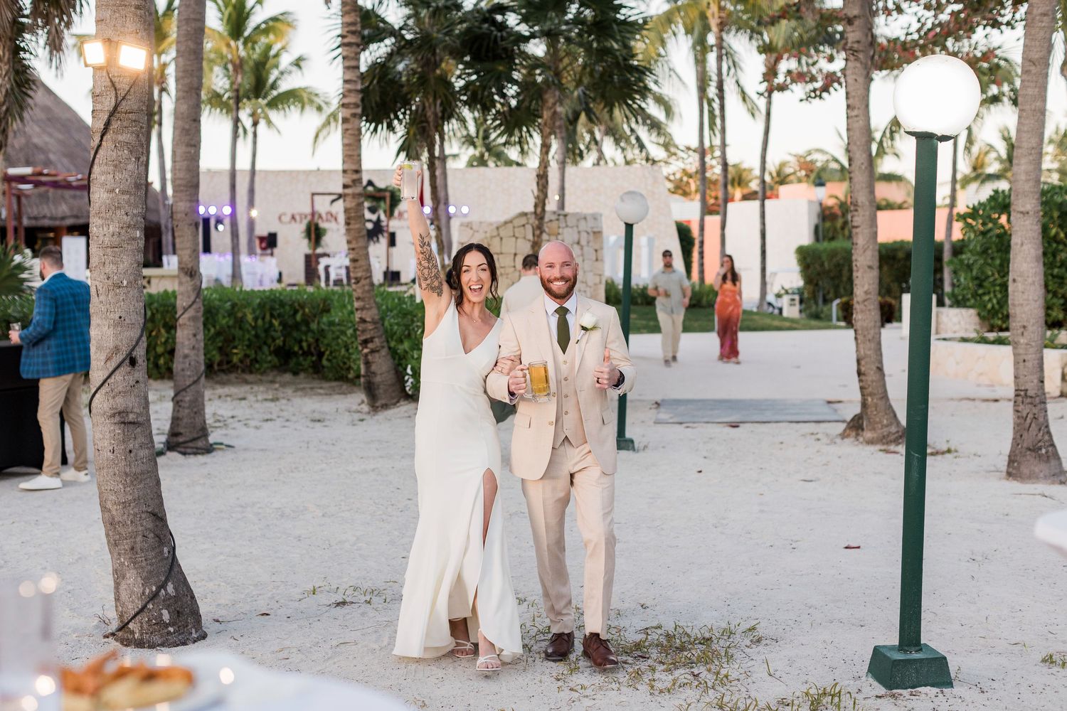 Bride and groom walking on sandy beach at outdoor tropical wedding reception, holding drinks and smiling among palm trees.