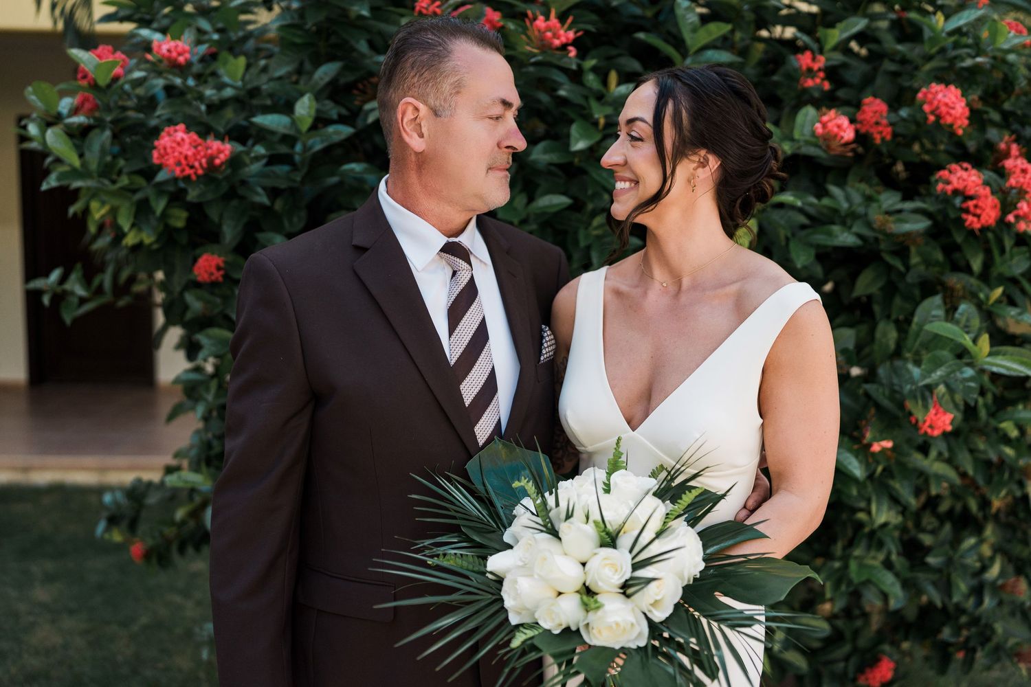 Bride and groom smiling at each other outdoors, bride holding white tropical bouquet, red flowers in background.
