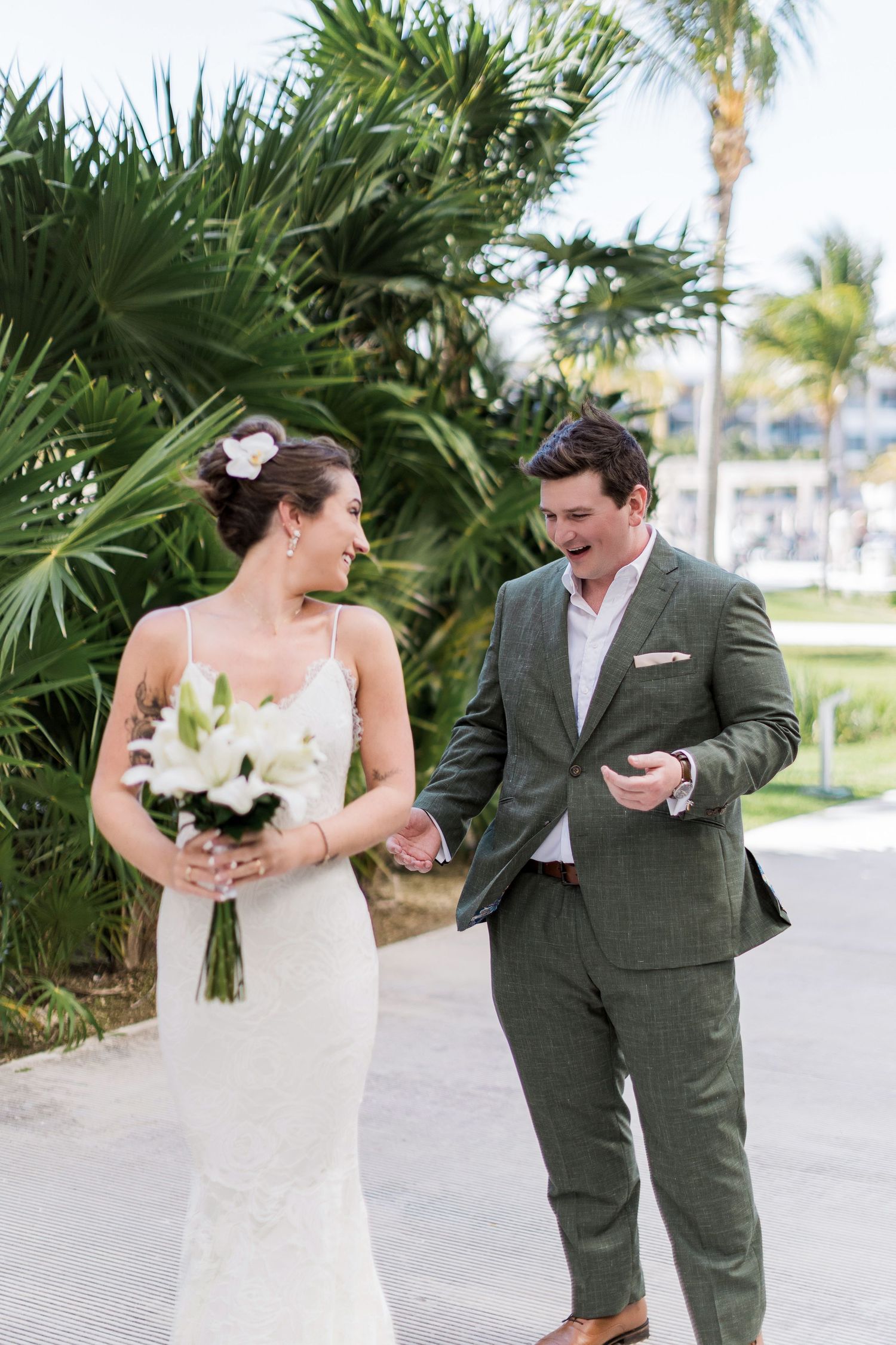 Bride in white gown holding lily bouquet smiles at groom in green suit during outdoor tropical wedding first look.