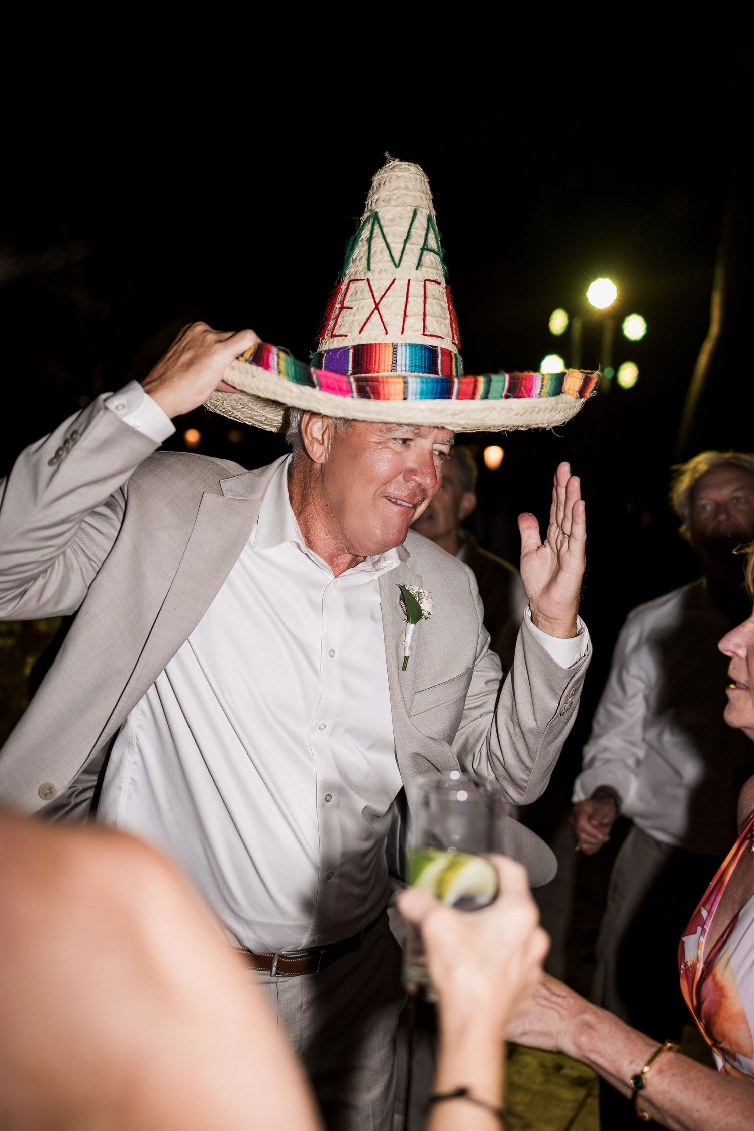 Man wearing colorful sombrero hat dancing at outdoor wedding reception at night.