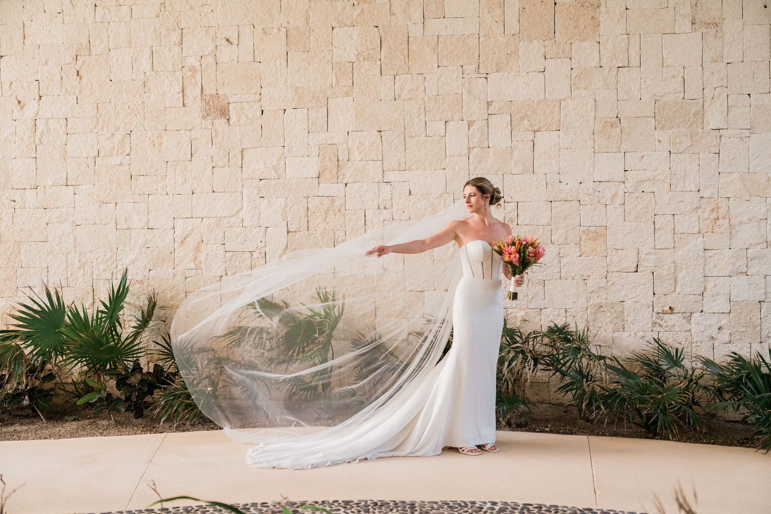 Bride in sleek white gown tosses flowing veil against a stone wall with tropical greenery backdrop.