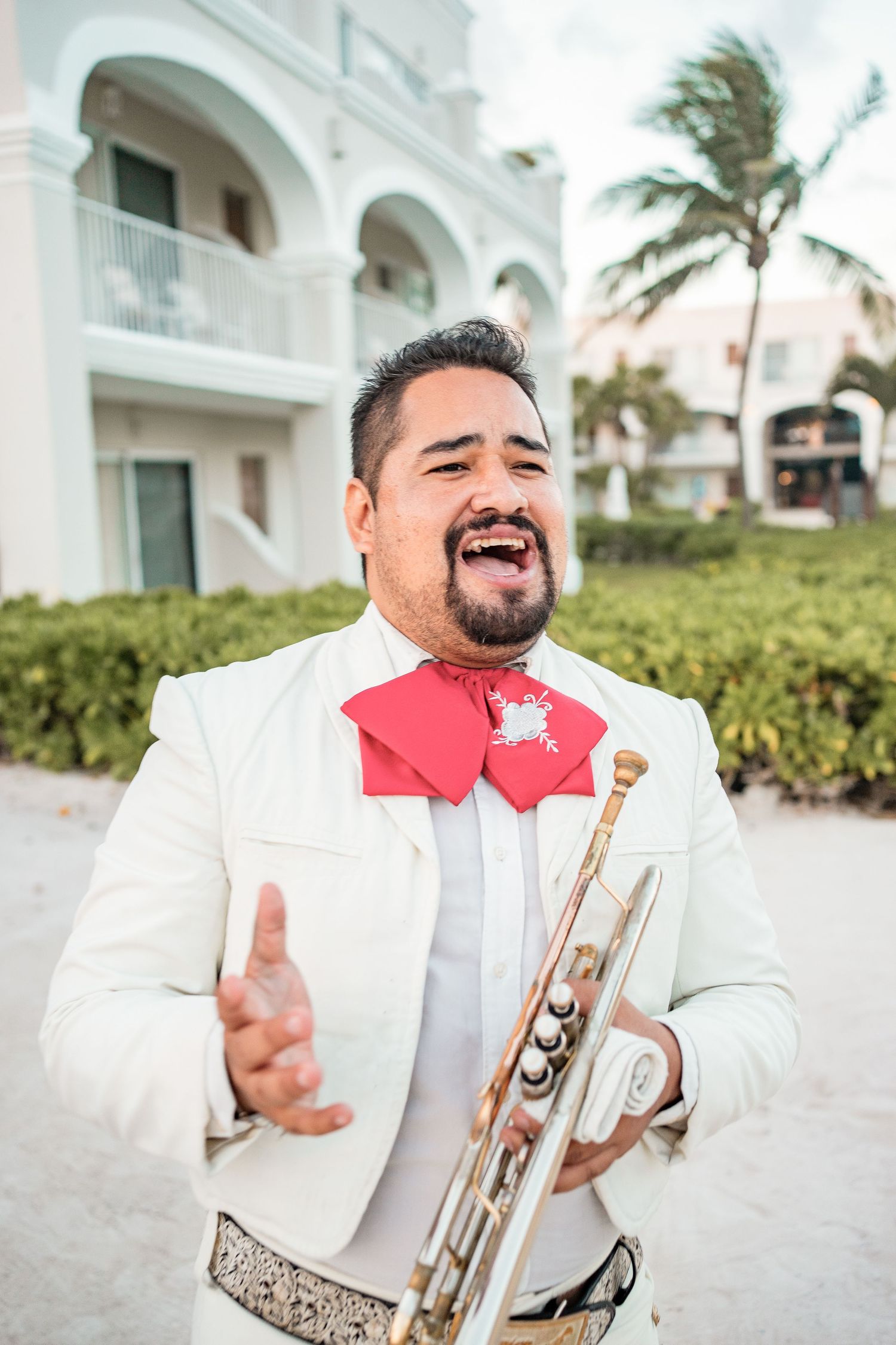 Mariachi trumpet player in white suit and red bow tie performs at tropical beach wedding.