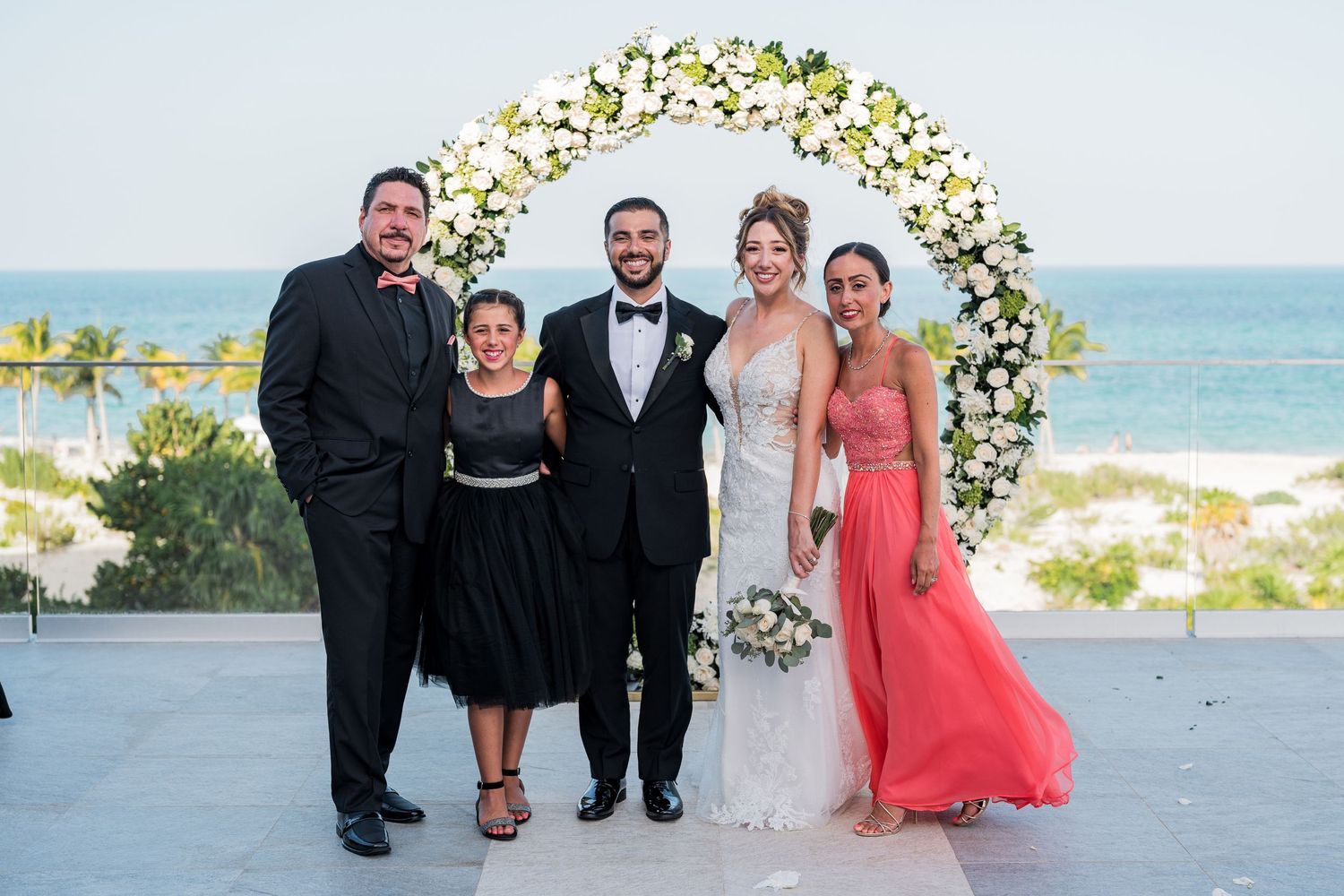 Bride and groom pose with family members under white floral arch at beachside wedding.