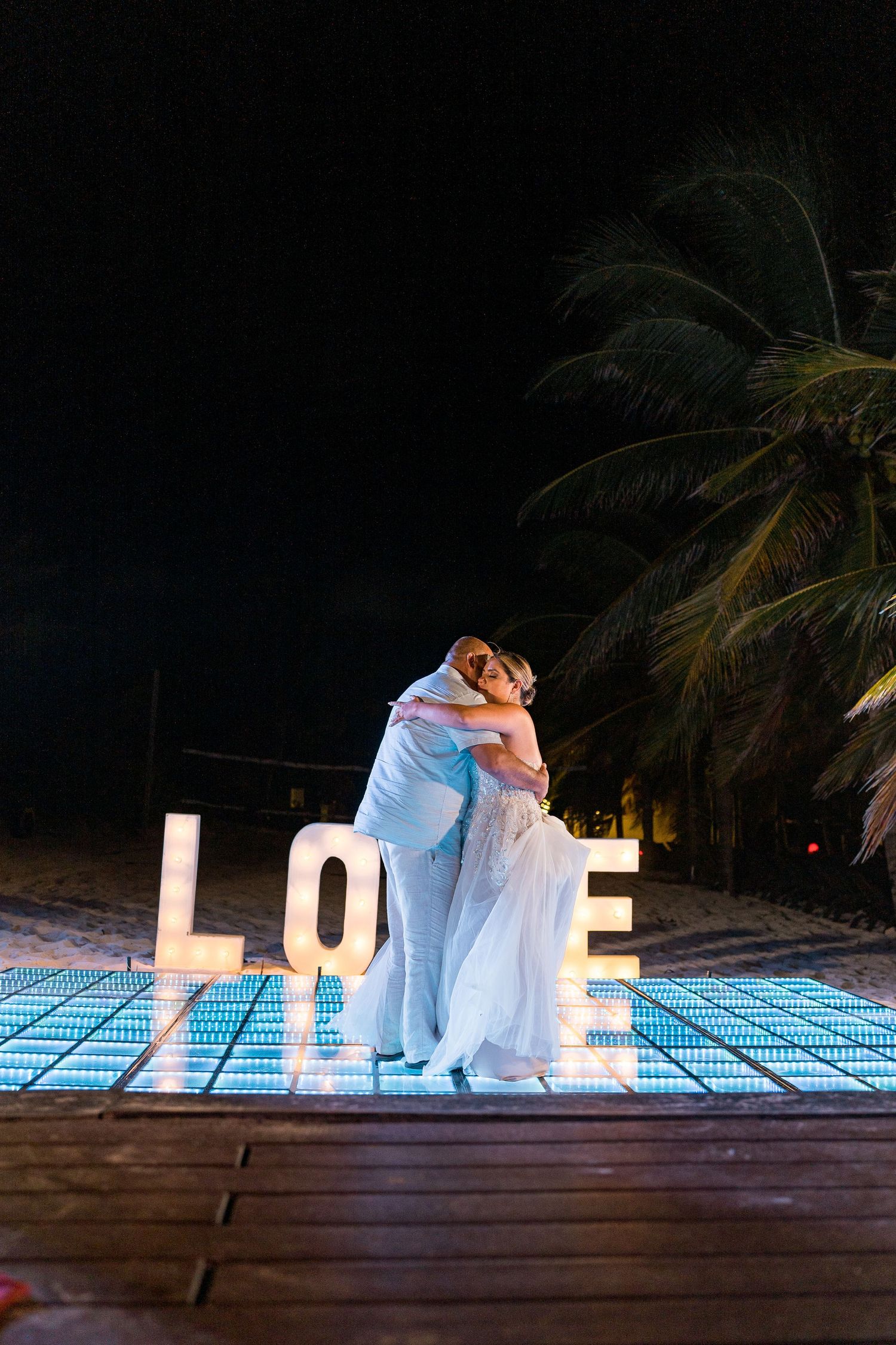 Bride and groom embracing on glowing dance floor with large LOVE marquee letters at night.