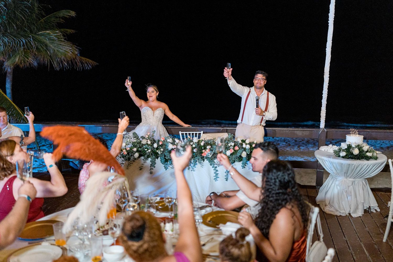 Bride and groom celebrating with raised hands at tropical beach wedding reception with guests cheering.