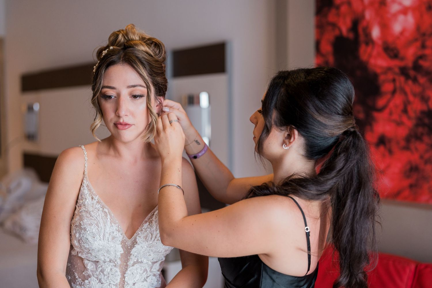 Bridesmaid helping bride put on earrings while wearing beaded lace wedding gown before ceremony.
