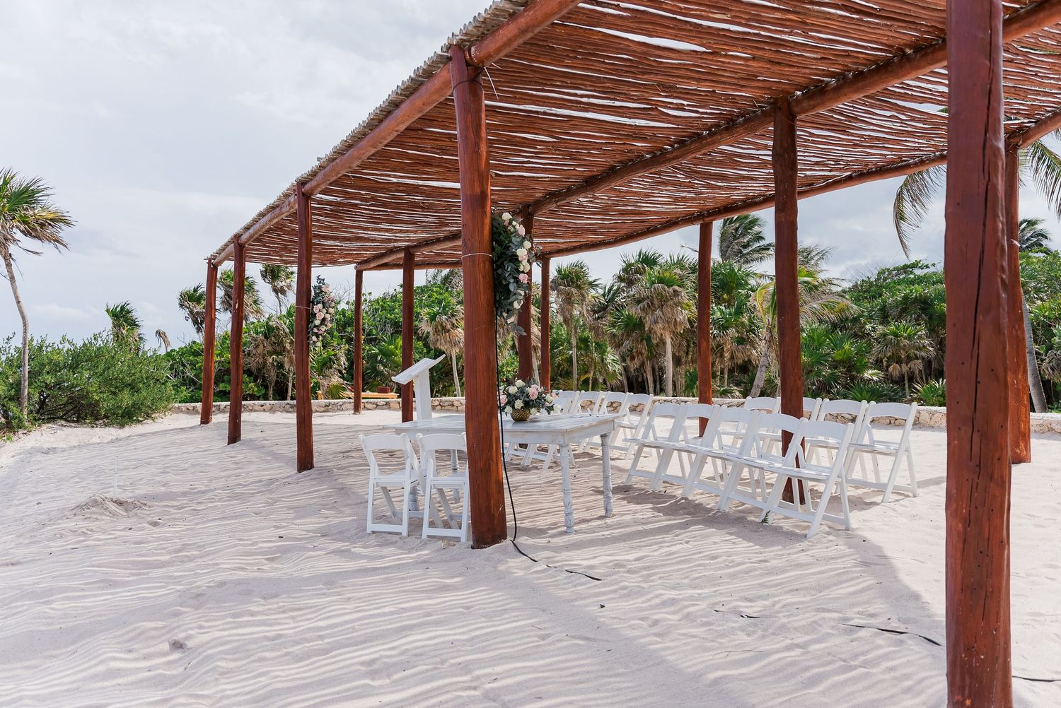 White folding chairs set up for outdoor beach wedding ceremony under rustic wooden pergola structure.
