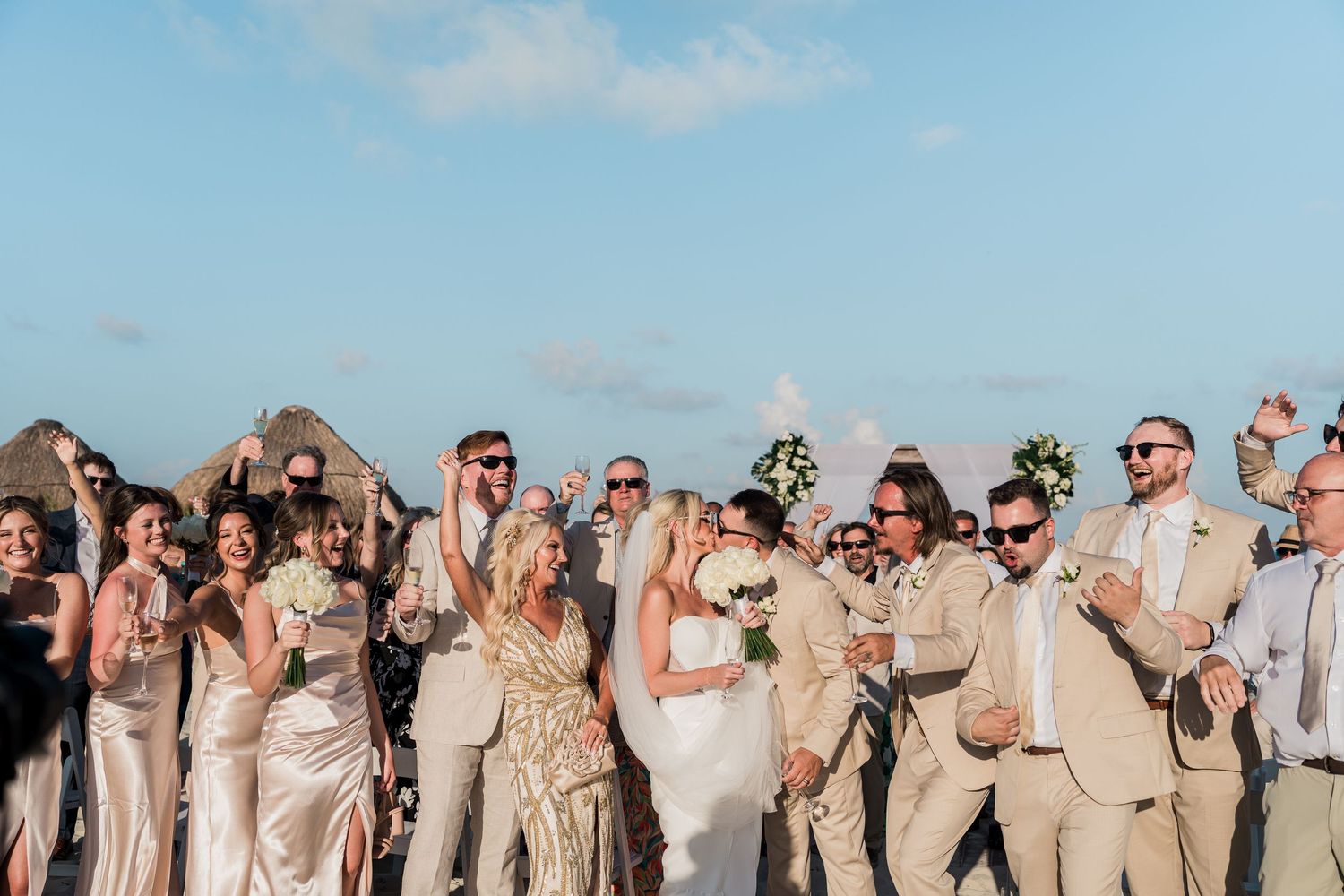 Joyful newlyweds celebrate with wedding party on beach, bride holding white bouquet, guests cheering around them.