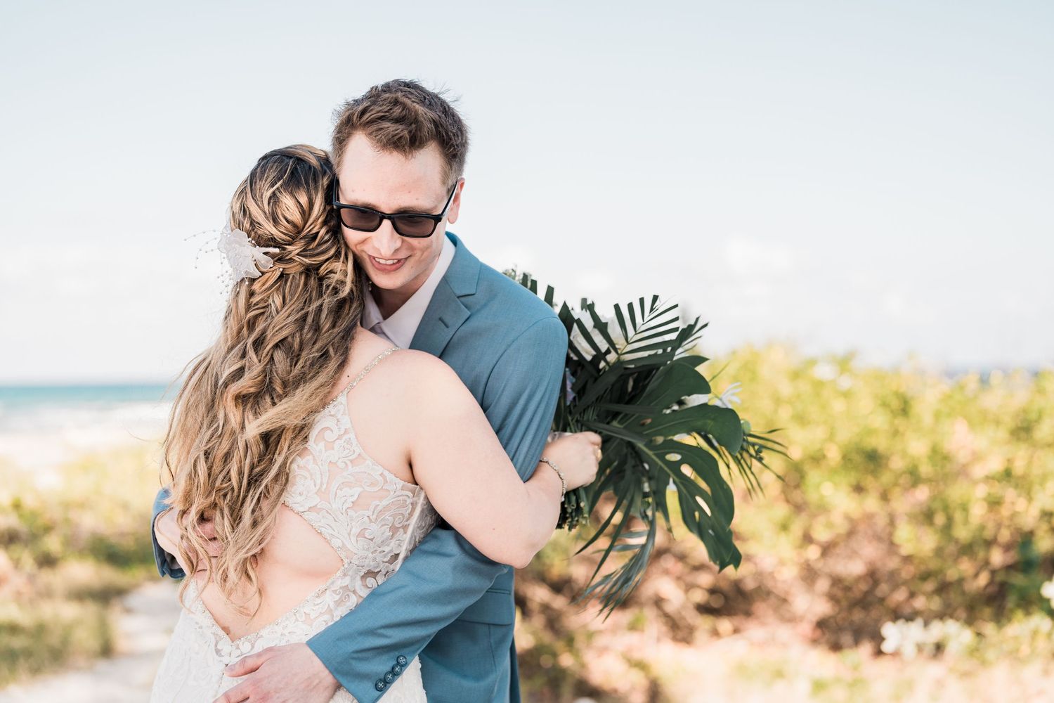 Groom embraces bride holding tropical palm leaf bouquet during emotional first look on beach.