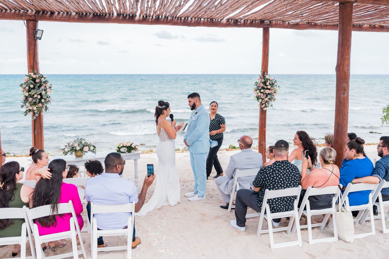 Beach wedding ceremony with couple at the altar, ocean backdrop, guests seated in white chairs.