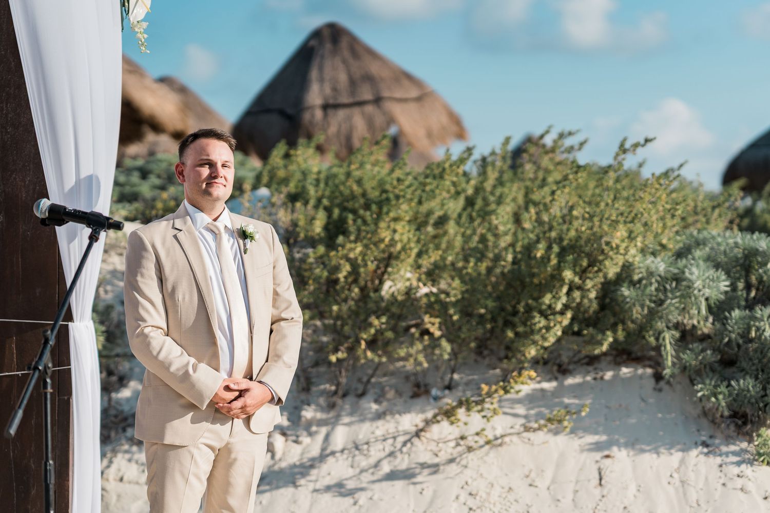 Groom in beige suit stands at beach wedding altar with sandy dunes and blue sky behind.