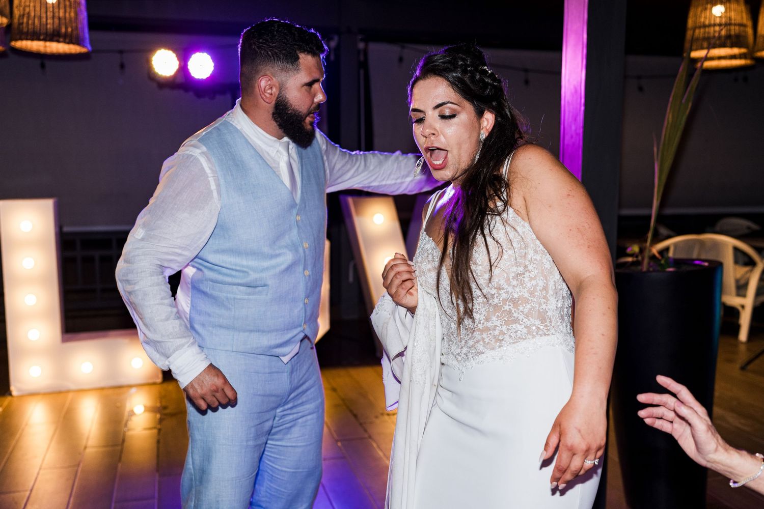 Bride and groom dancing energetically at their wedding reception with illuminated letter decor in the background.