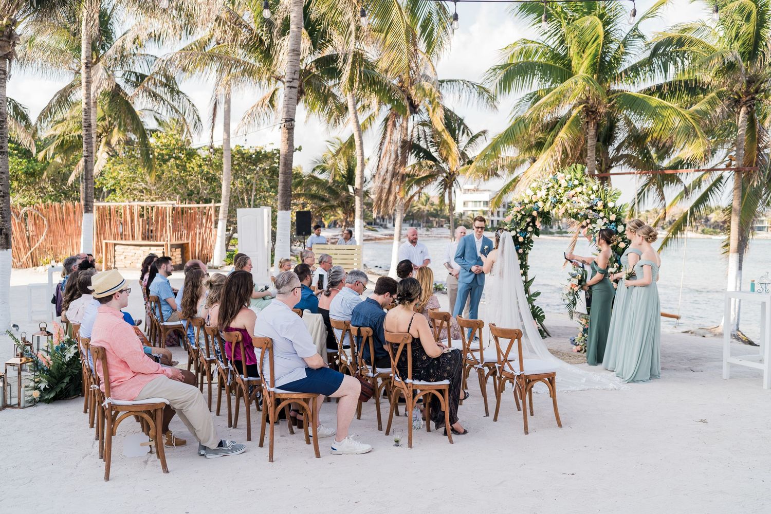 Outdoor beach wedding ceremony with guests seated on wooden chairs surrounded by tropical palm trees.