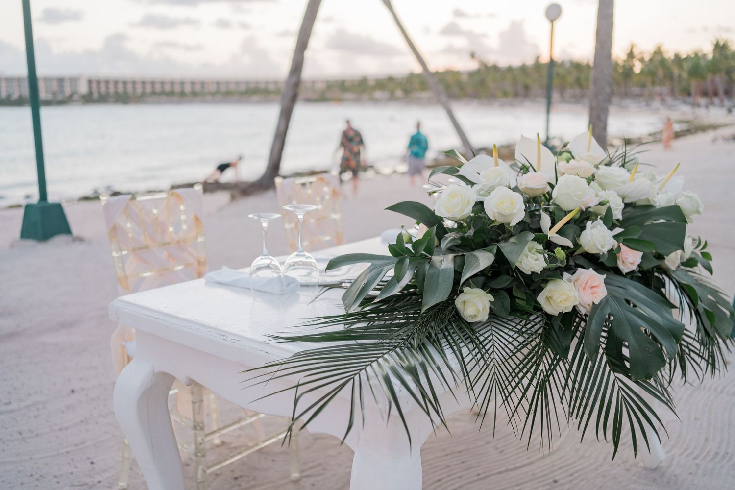 White floral arrangement with tropical leaves on beach wedding ceremony table at sunset.
