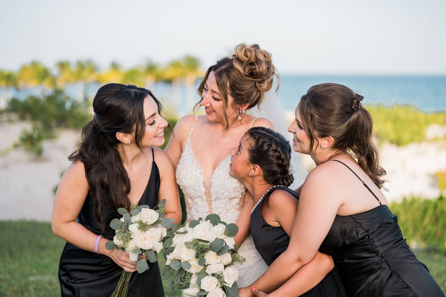 Bride and bridesmaids share a joyful moment together on a green lawn with ocean and palms behind.