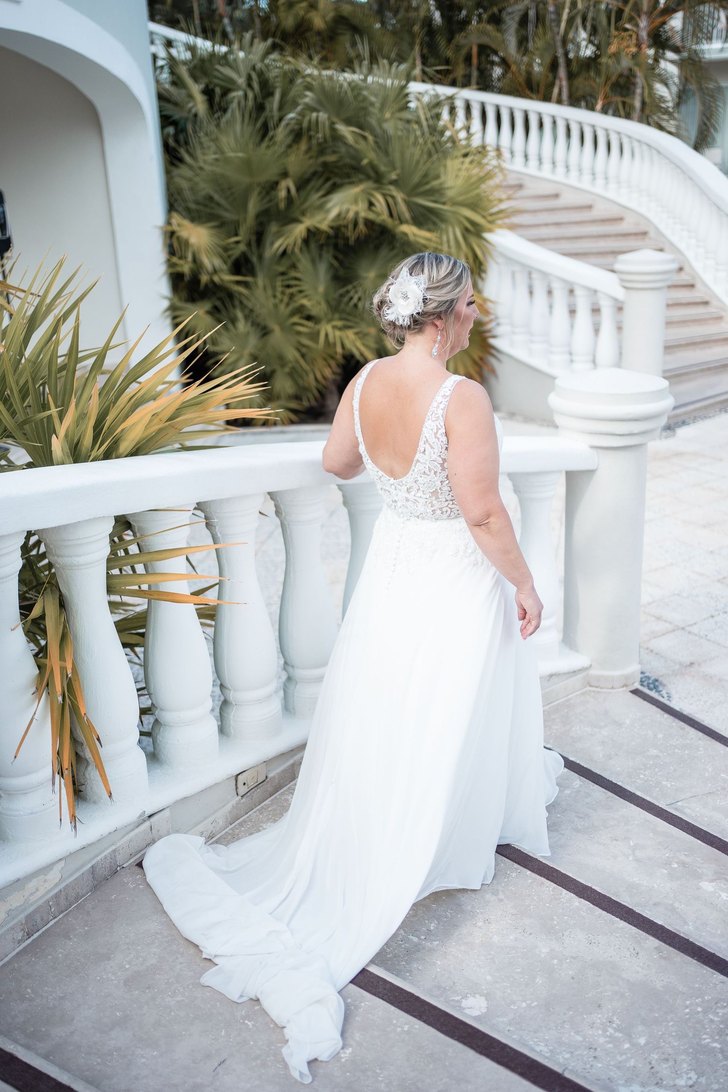 Bride in elegant backless lace wedding gown stands by white balustrade with tropical greenery backdrop.