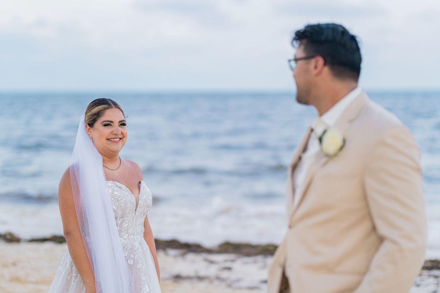 Bride in white lace wedding dress and veil smiling at groom in beige suit on a scenic ocean beach.