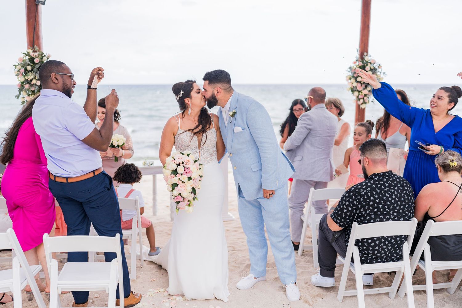 Bride and groom share romantic kiss during beach wedding recessional surrounded by cheering guests.