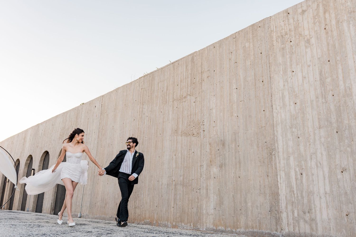 A joyful couple in wedding attire runs hand-in-hand along a modern concrete wall in an outdoor setting.