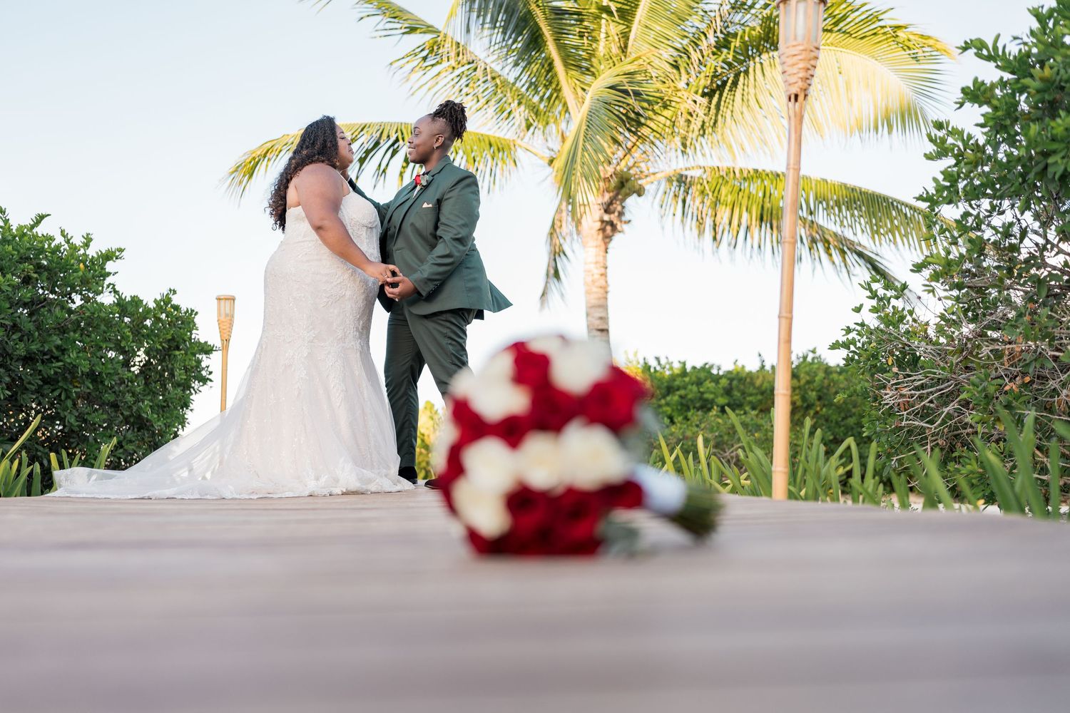 Bride and groom pose on wooden boardwalk with red rose bouquet in foreground.