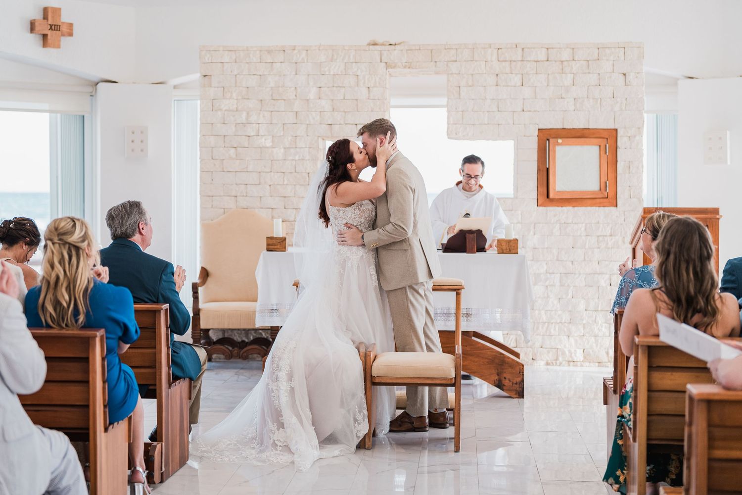 Bride and groom share their first kiss at the altar in a sunlit white chapel wedding ceremony.