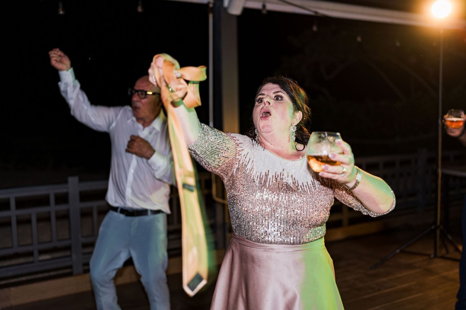 Lively wedding guest waves a tie while holding a drink on the outdoor dance floor.