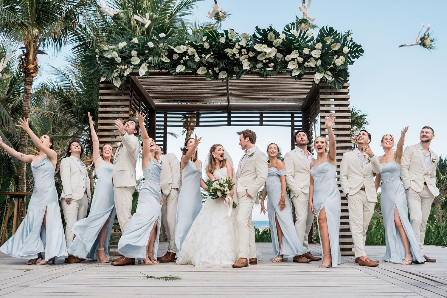 Wedding party poses joyfully under a floral arch at a tropical beach venue with palm trees.