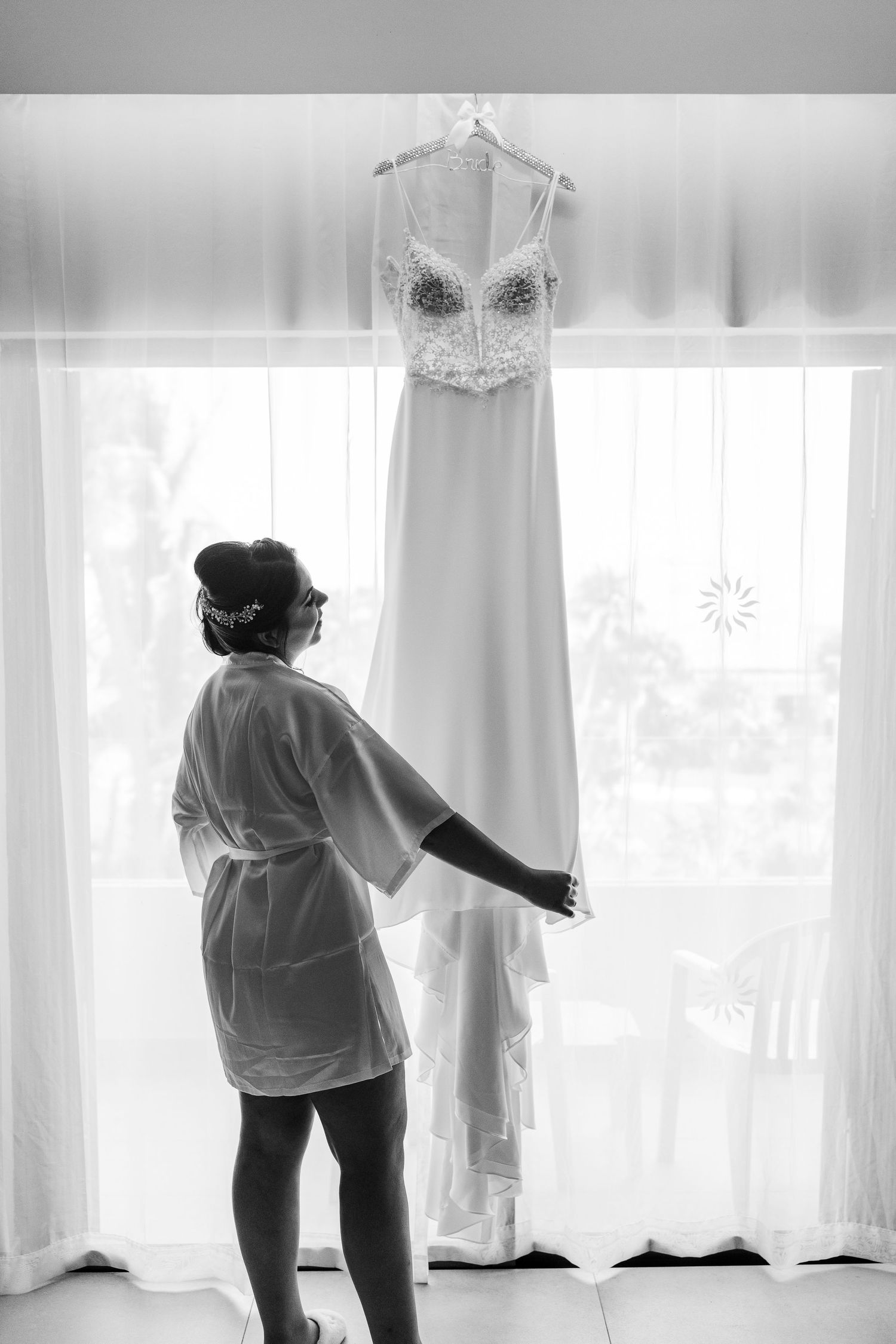 Black and white photo of bride in robe reaching for wedding dress hanging by window.