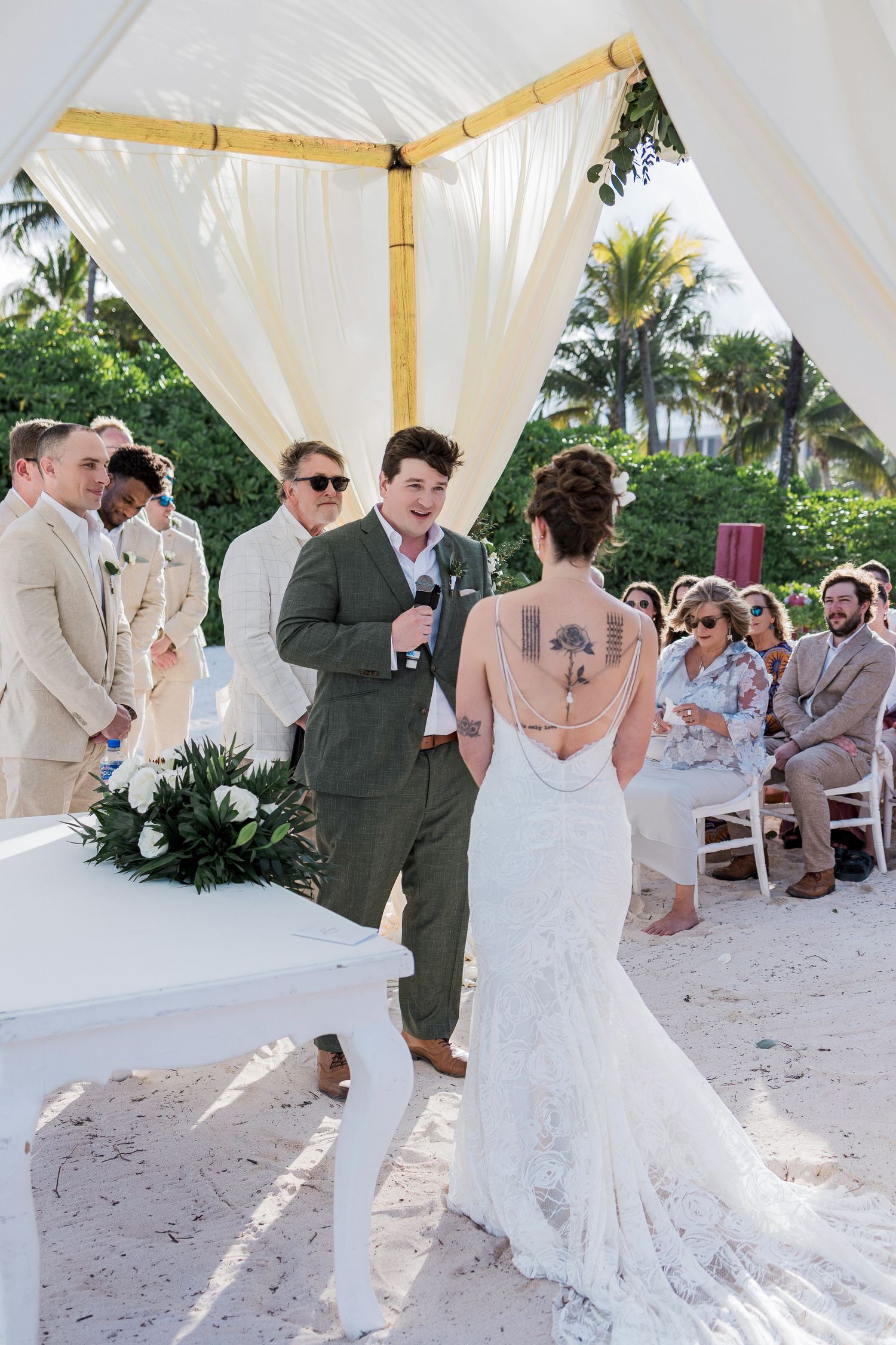 Groom reciting vows to bride in lace backless wedding dress at outdoor beach ceremony under white draped canopy.