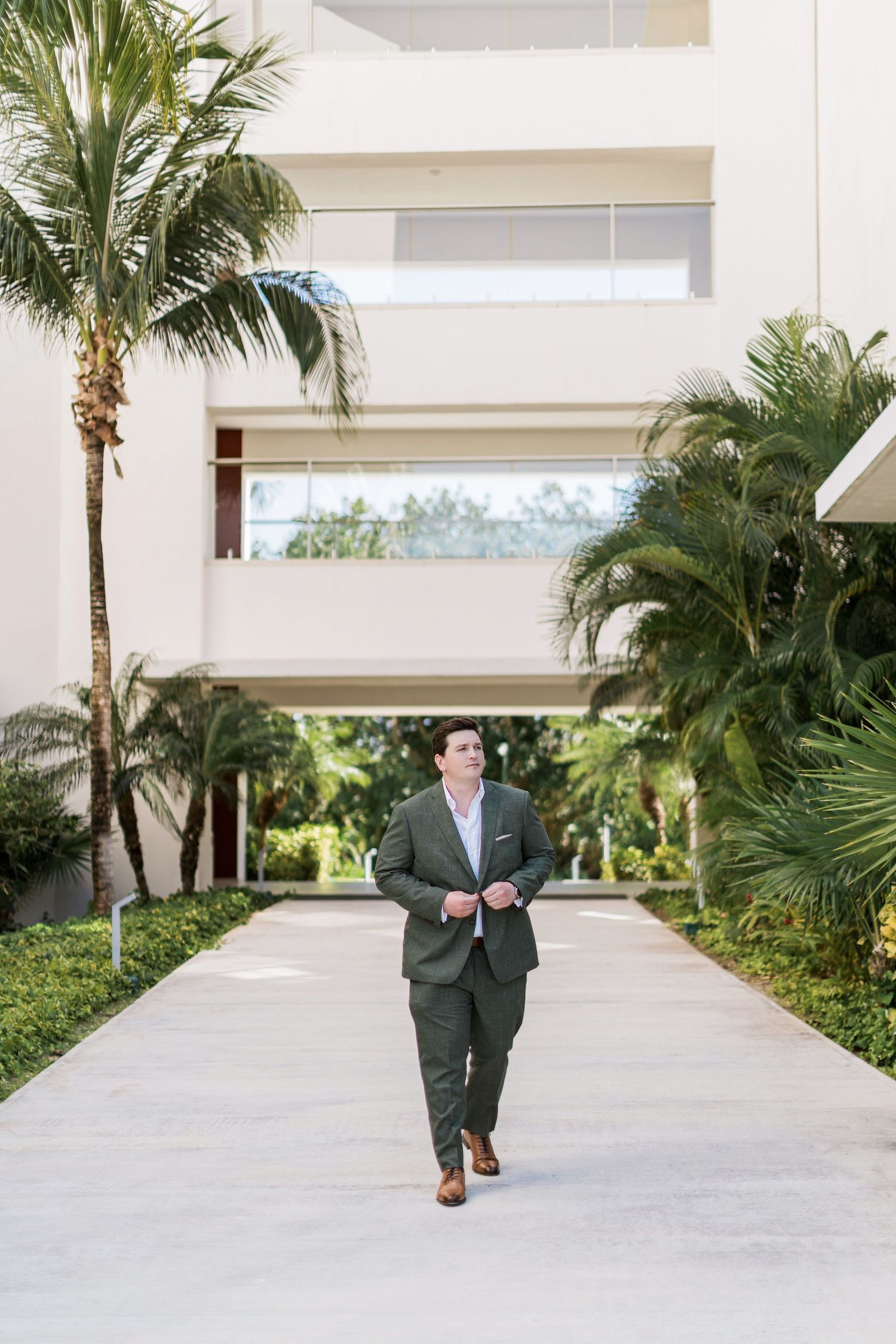 Man in green suit walking down a palm-lined pathway at a modern tropical resort building.