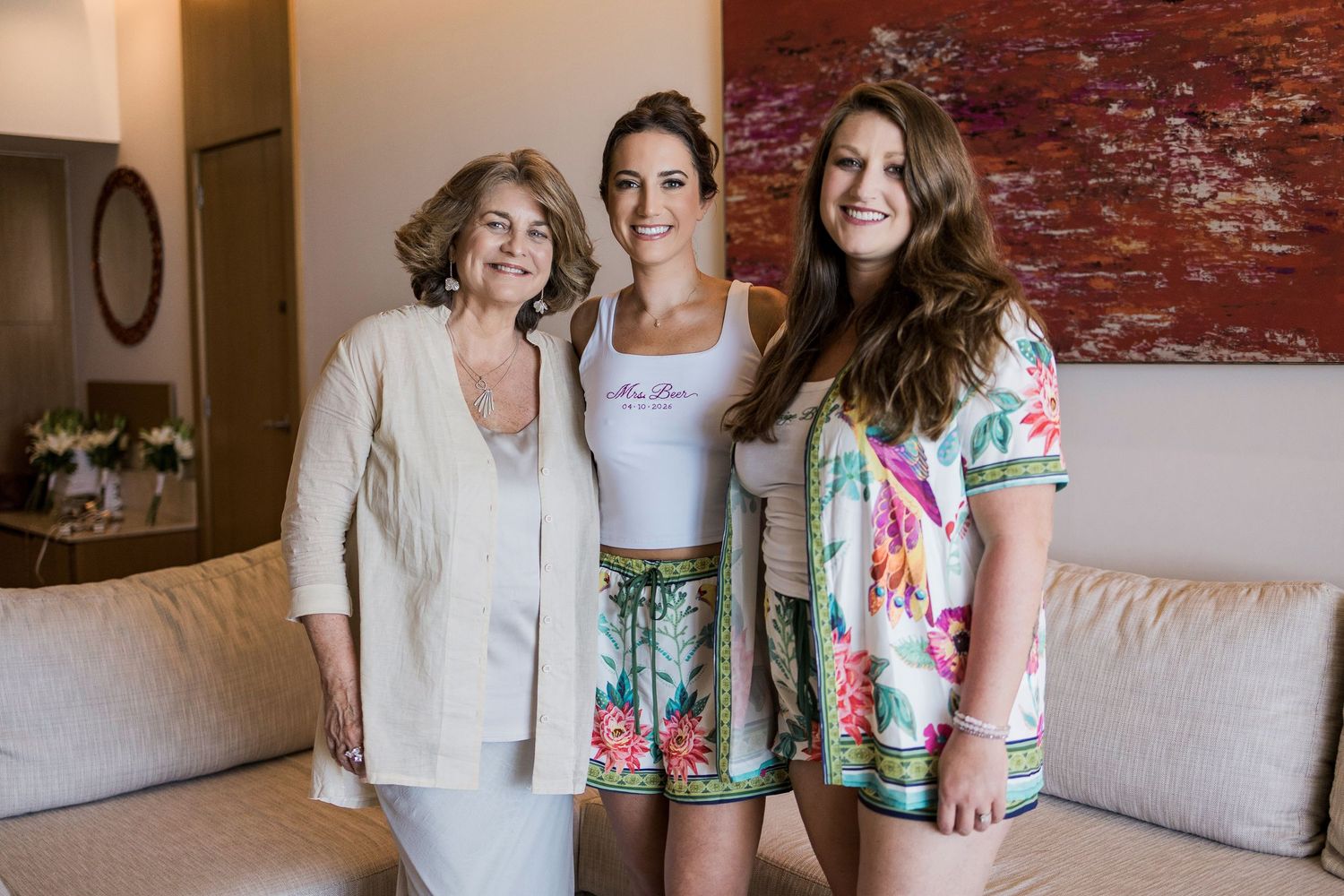 Three women smiling together indoors, dressed in casual summer outfits, posing in a bright modern room.