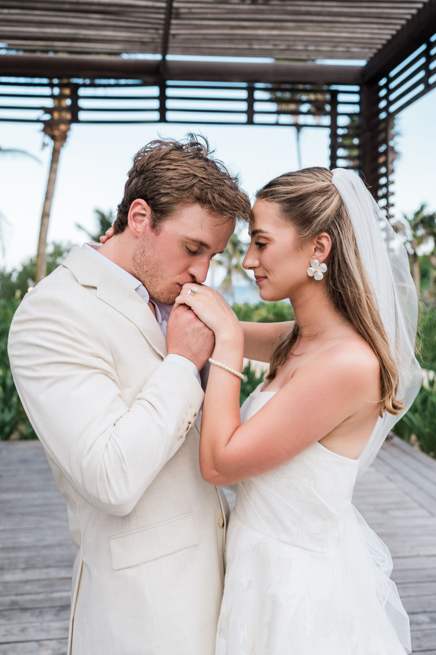 Groom in ivory suit tenderly kisses bride's hand during romantic outdoor wedding portrait at tropical resort.