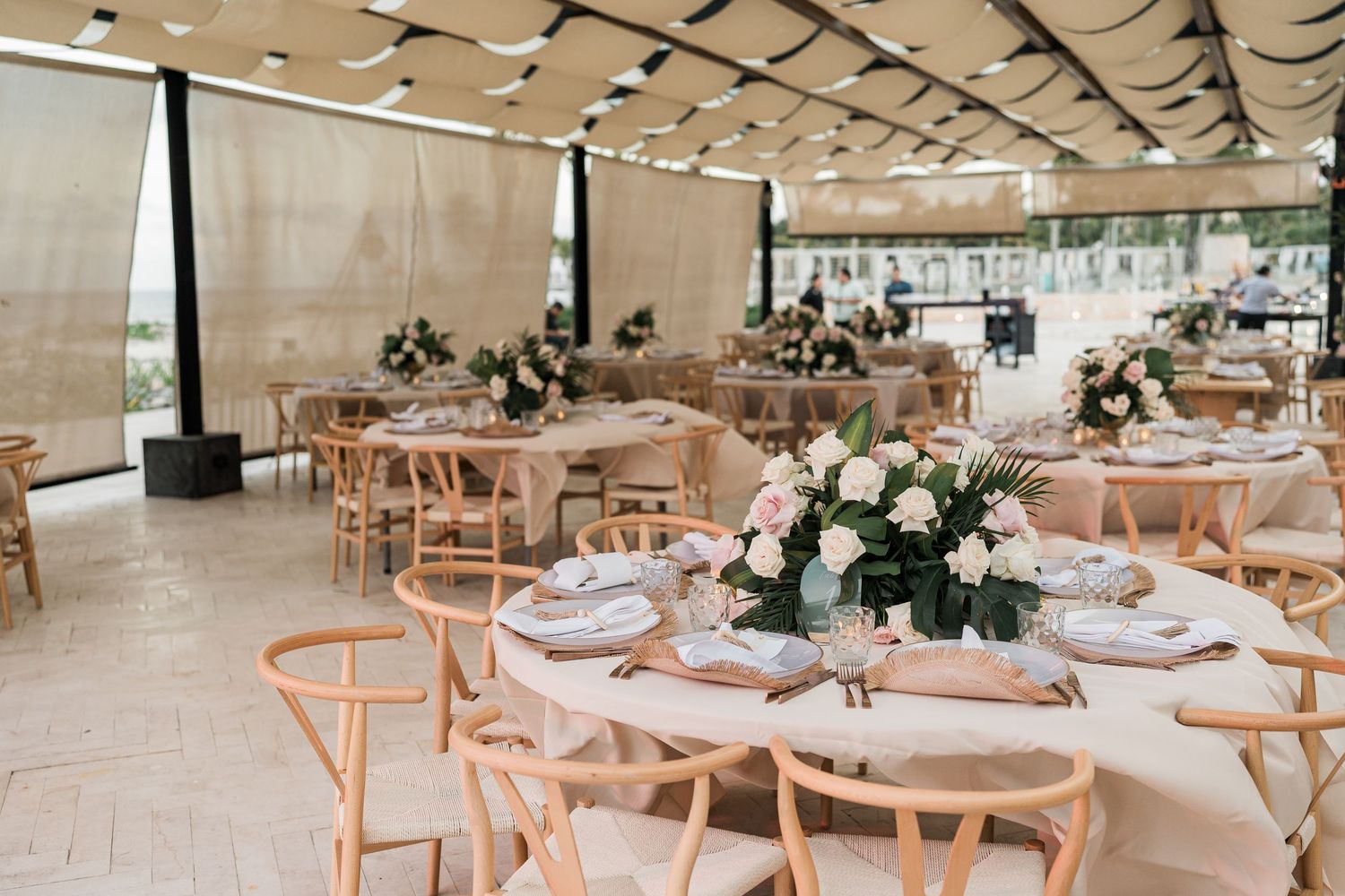Elegant wedding reception venue with wishbone chairs, round tables, floral centerpieces, and string lights under a draped canopy.
