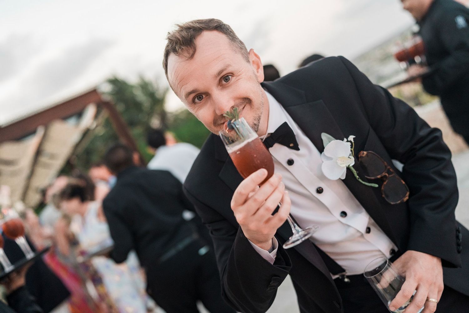 Man in tuxedo with bow tie smiling while holding a cocktail glass at an outdoor wedding reception.