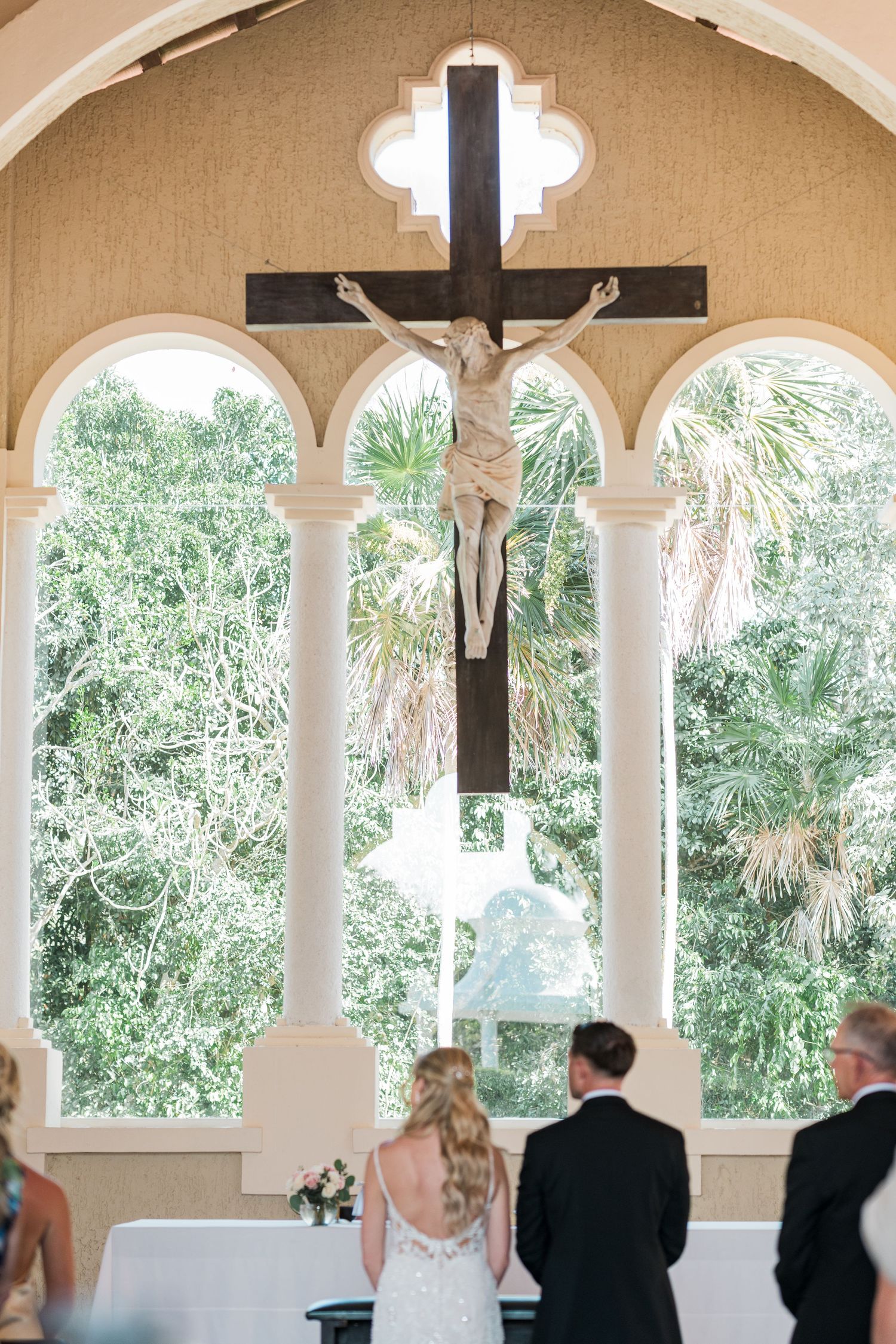 Bride and groom stand before a crucifix at a chapel altar with arched windows and lush greenery backdrop.