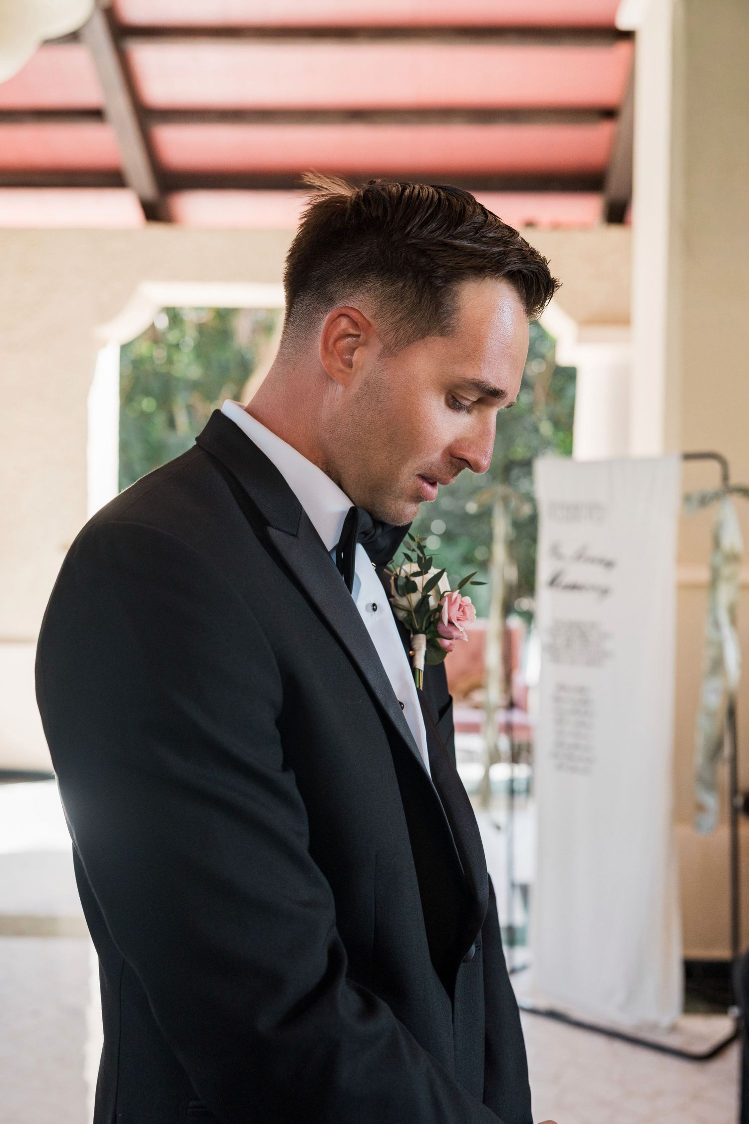 Groom in black tuxedo with pink boutonniere looking down thoughtfully at outdoor wedding venue.
